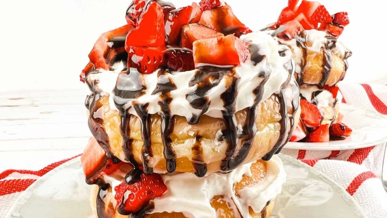 A stack of donuts topped with whipped cream, chocolate drizzle, and fresh strawberry pieces on a white plate.