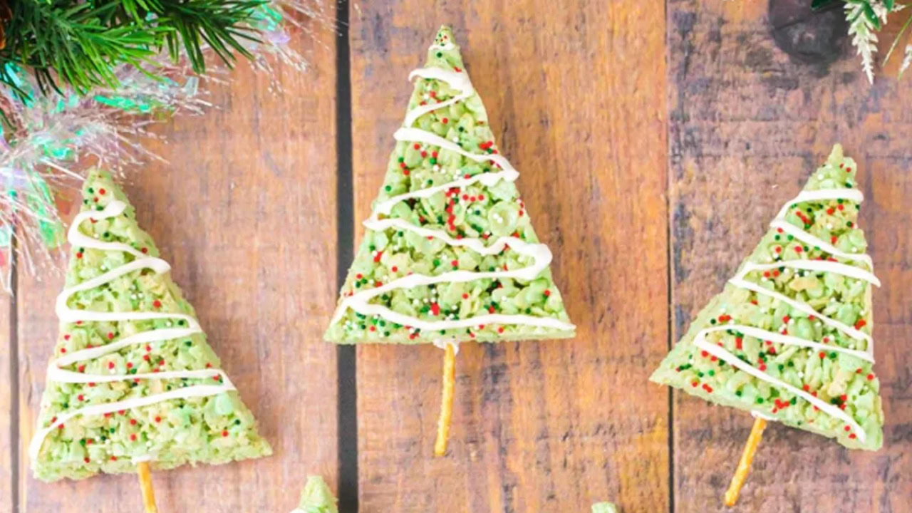 Three Christmas tree-shaped rice crispy treats with green coloring, white icing, and sprinkles are arranged on a wooden surface, each with a pretzel stick as the trunk.