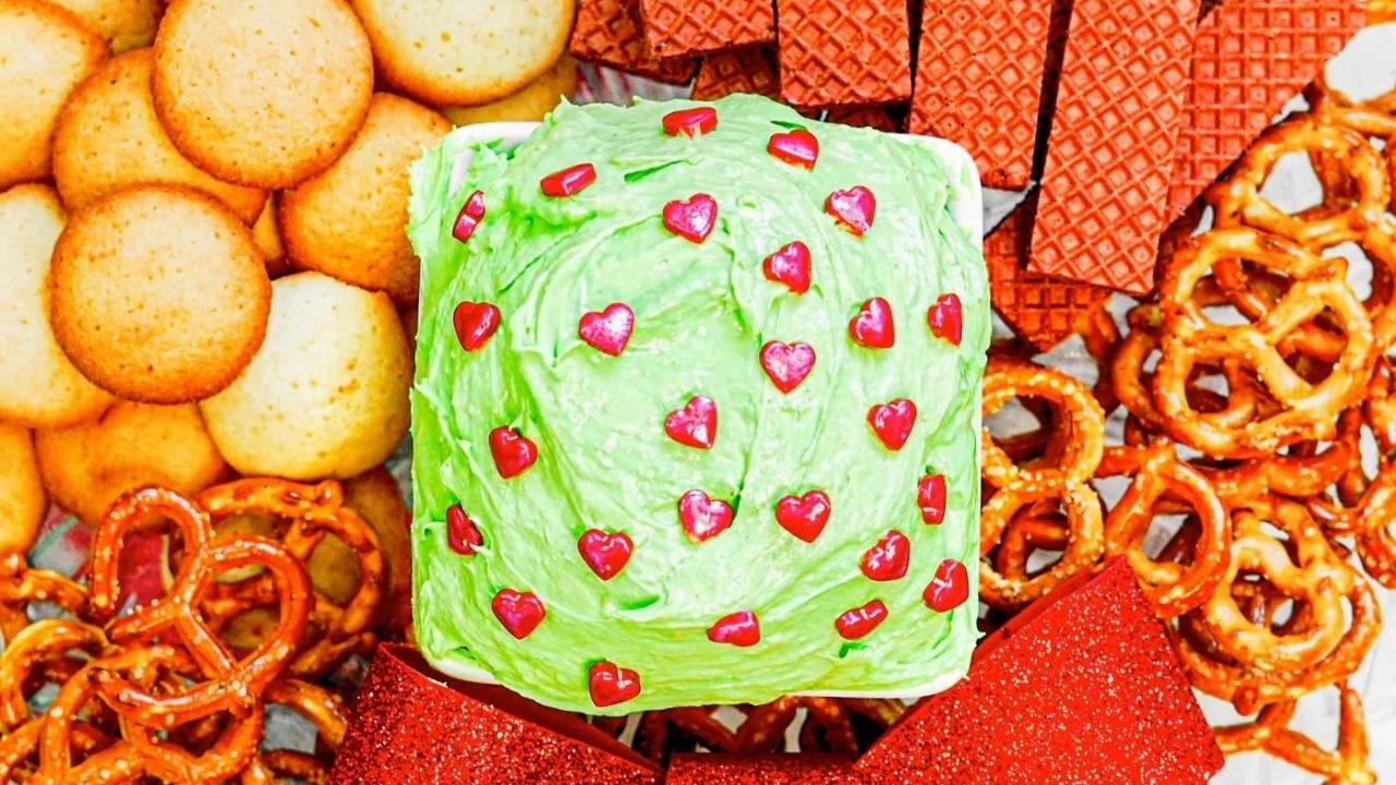 A bowl of green dip with red heart sprinkles is surrounded by vanilla cookies, red wafer cookies, and pretzels.
