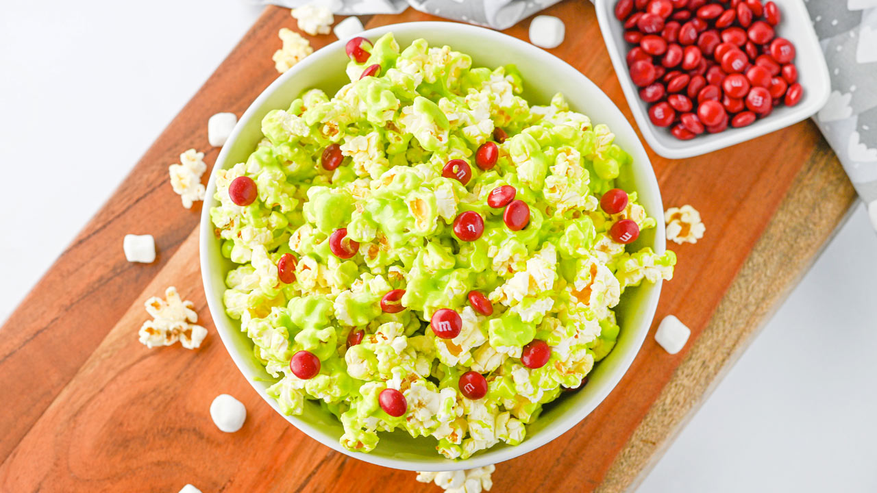 A bowl of green popcorn topped with red candy sits on a wooden surface, with extra popcorn, marshmallows, and a dish of red candies nearby.