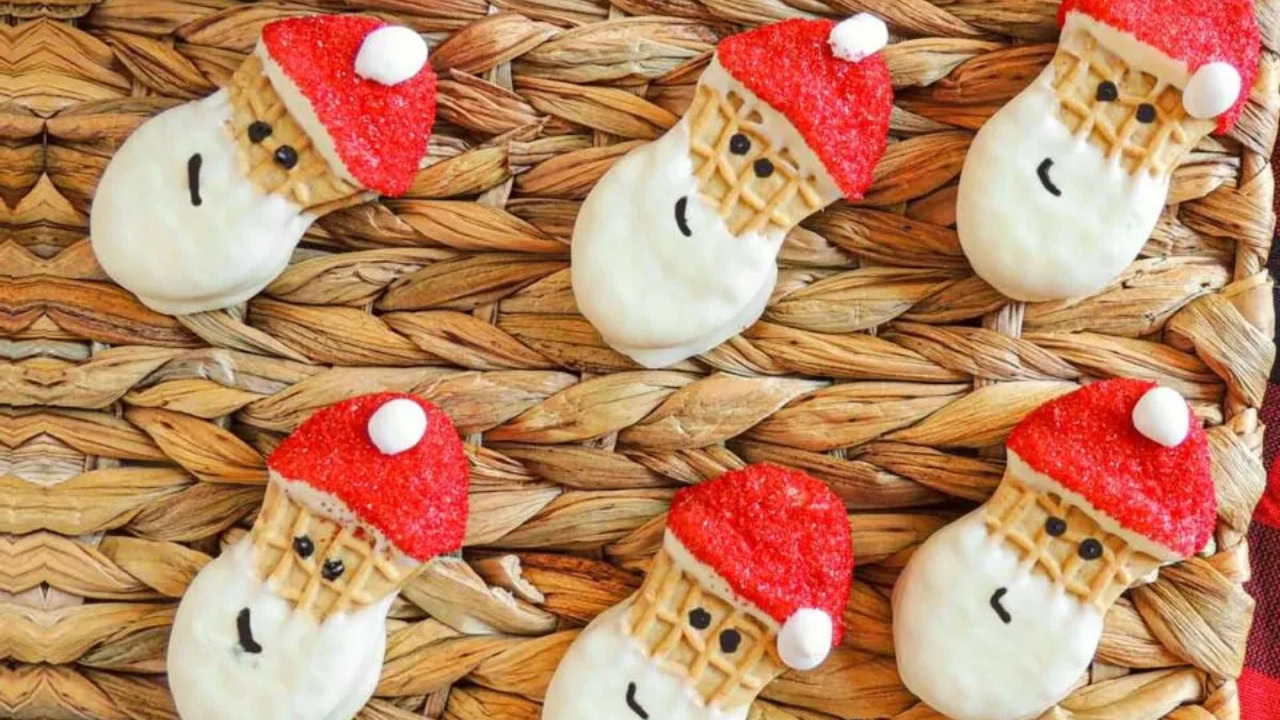 Six cookies decorated to look like Santa Claus with red hats and white beards are arranged on a woven placemat.