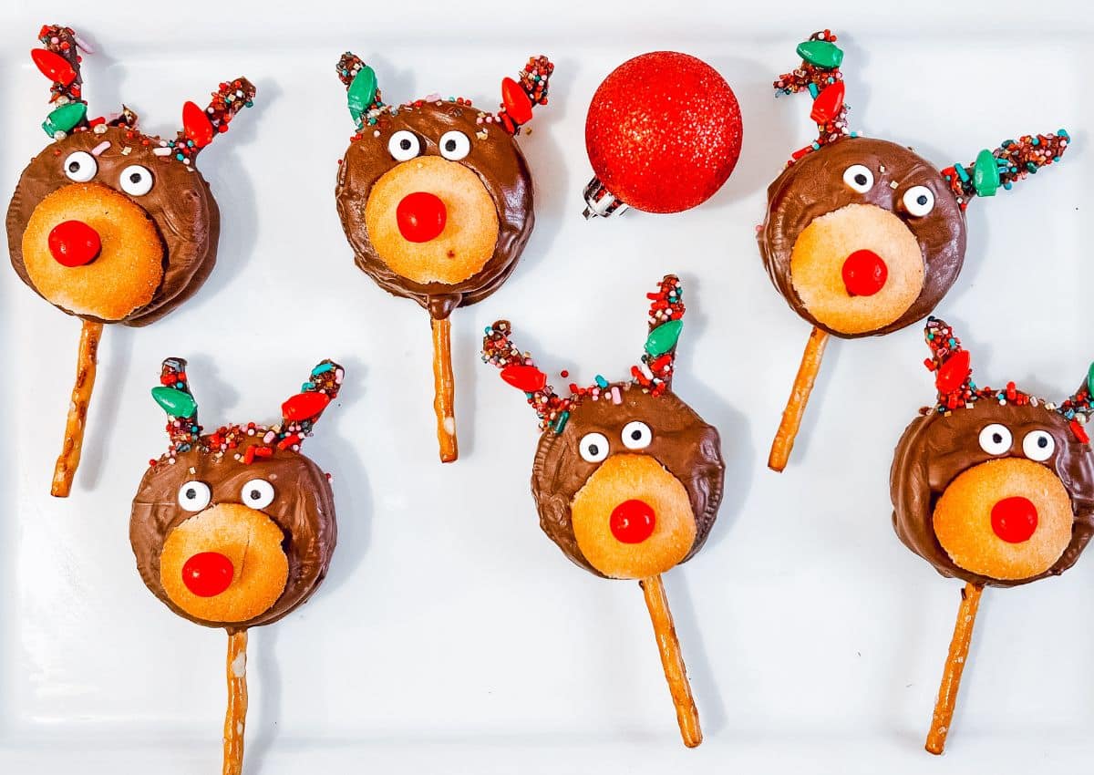 Six reindeer-shaped chocolate treats with pretzel antlers, candy eyes and noses, are arranged around a red ornament on a white surface.