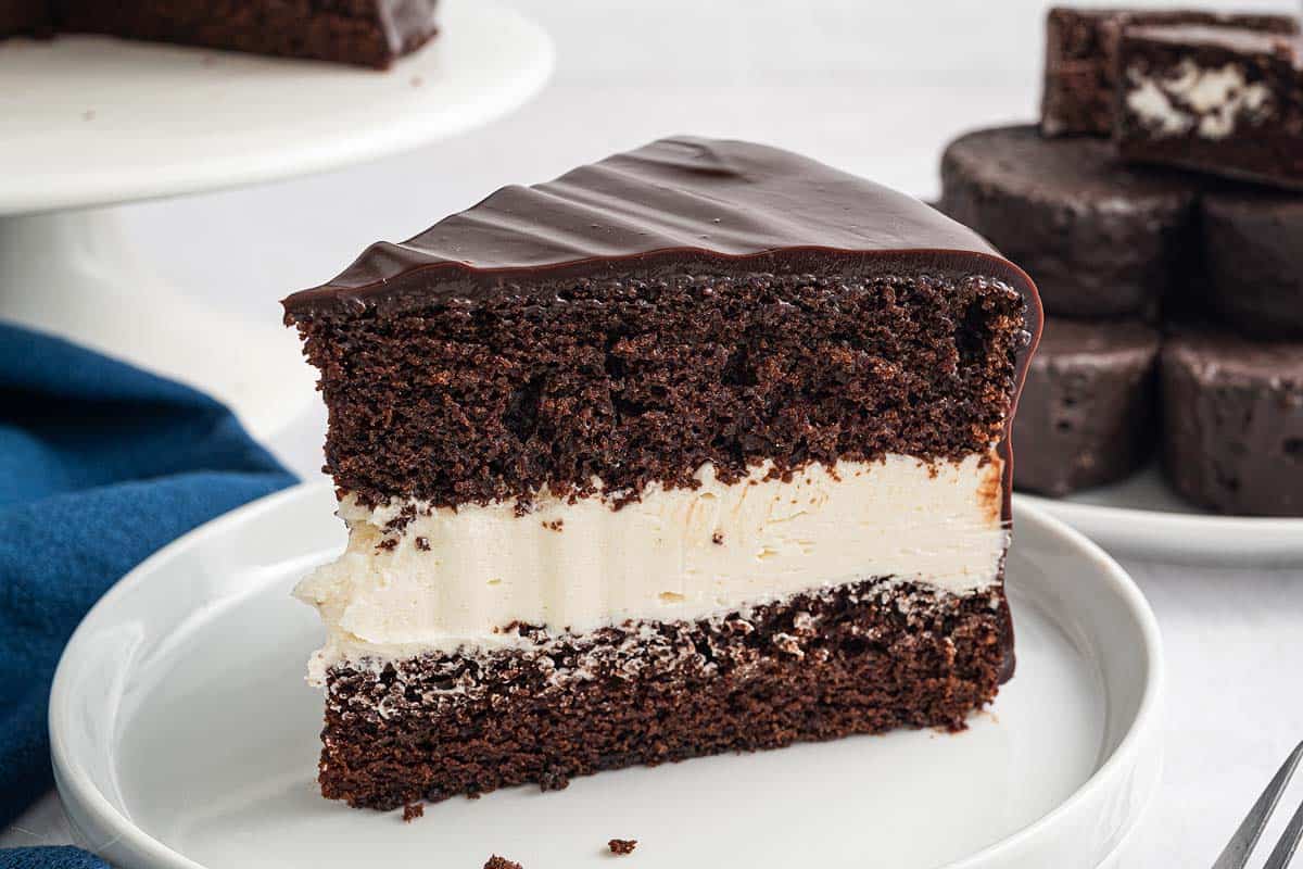 A slice of chocolate cake with a thick layer of white cream filling sits on a white plate; more cake pieces are visible in the background.