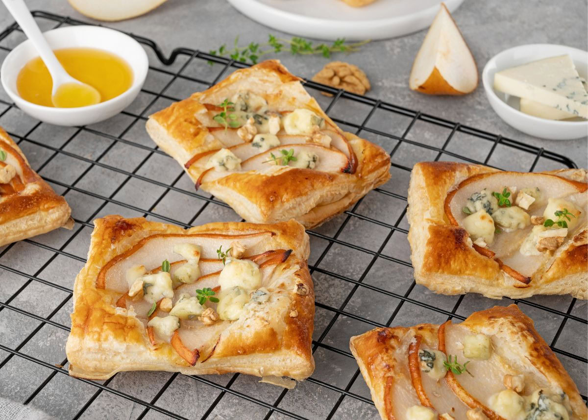 Square puff pastries topped with pear slices, blue cheese, and herbs are cooling on a wire rack; honey and cheese are in small bowls nearby.