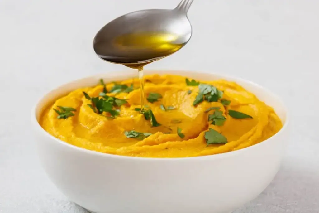 A spoon drizzles oil over a bowl of orange-colored dip garnished with chopped herbs.