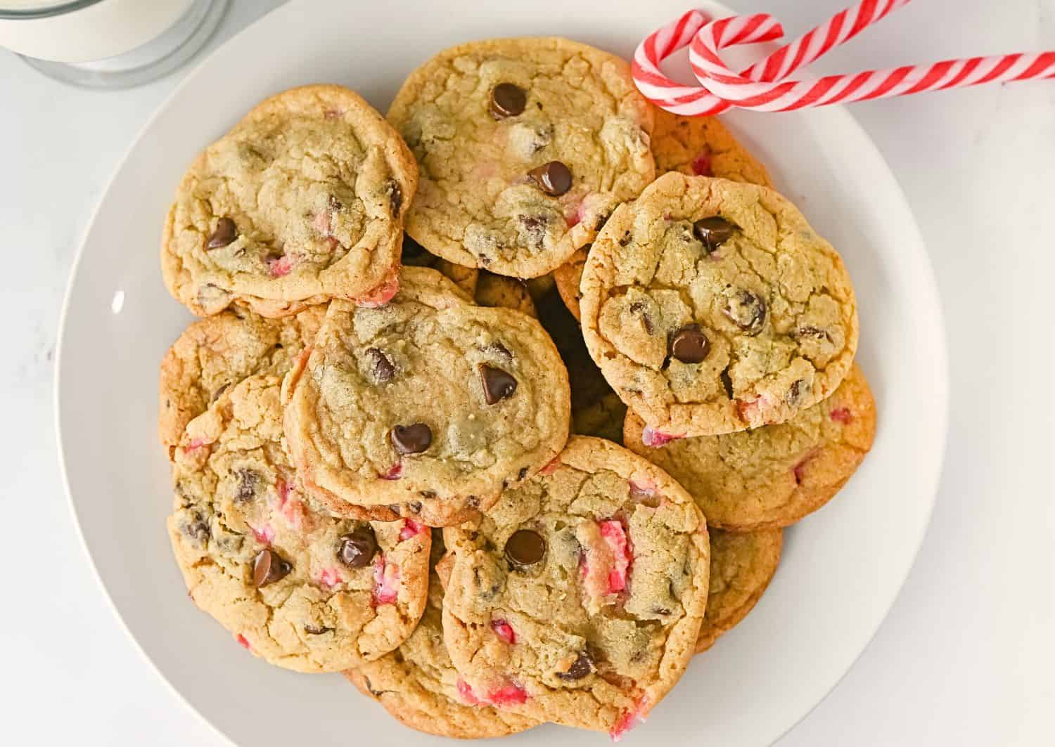 A plate of chocolate chip cookies with visible red candy pieces, next to two candy canes and a glass of milk.