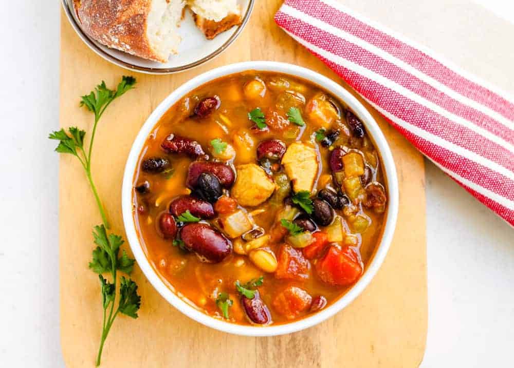 A bowl of mixed bean and vegetable stew on a wooden board, garnished with fresh parsley, with a slice of bread and a red-striped napkin nearby.