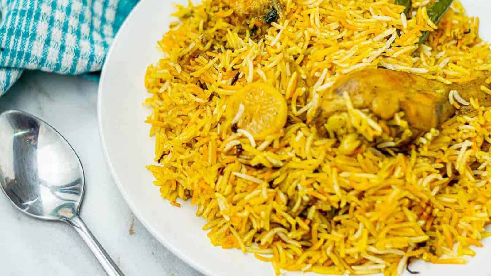 A plate of chicken biryani with yellow rice and chicken pieces, next to a silver spoon and a blue-checkered cloth.
