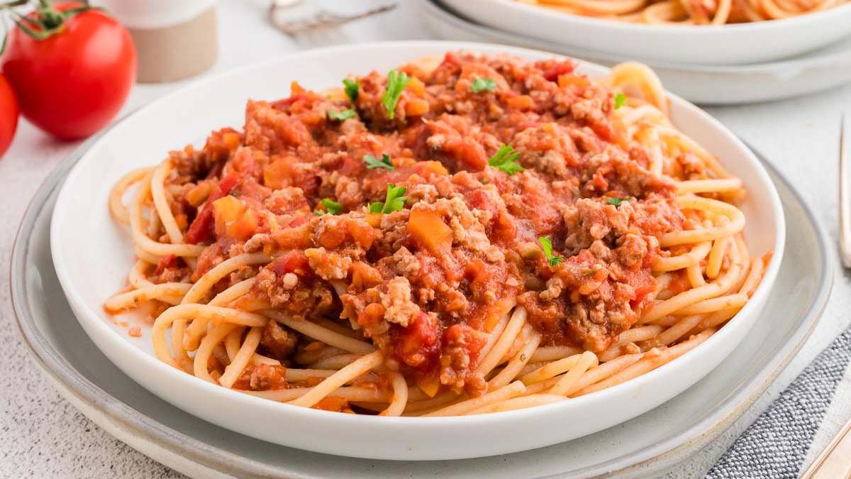A plate of spaghetti topped with ground meat and tomato sauce, garnished with chopped herbs, sits on a white plate with another plate of spaghetti in the background.