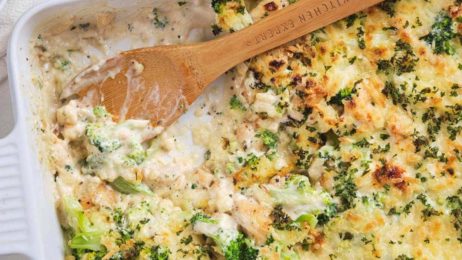 A baked broccoli casserole with a creamy sauce, partially served, in a white dish with a wooden spoon resting inside.