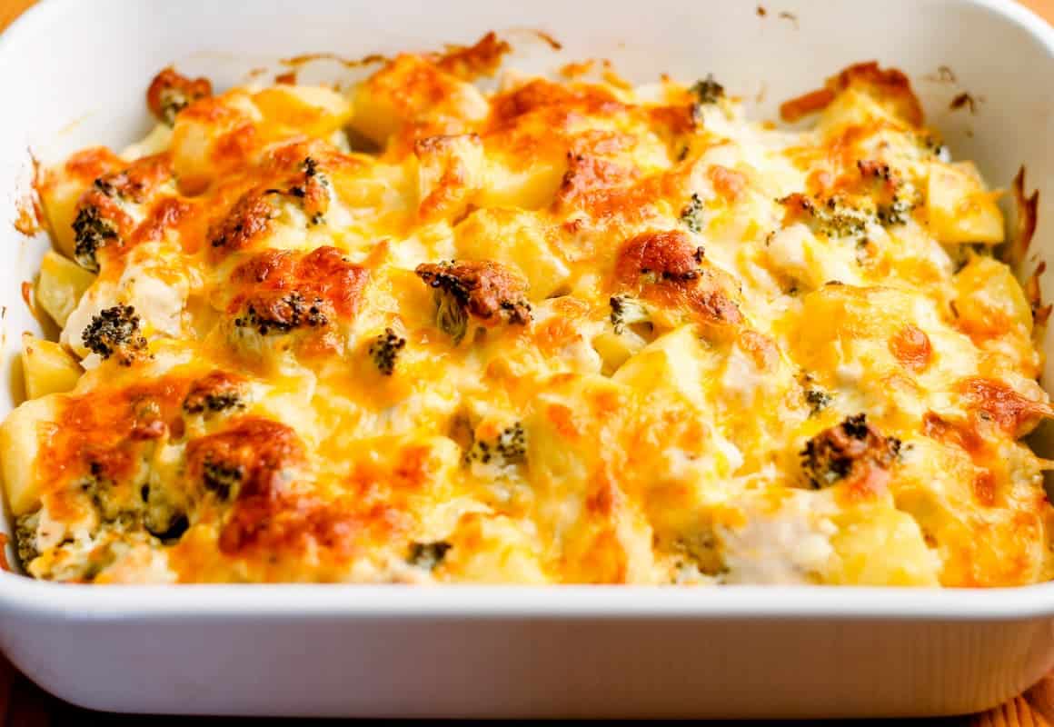 A baked casserole in a white dish, featuring browned melted cheese on top of vegetables, likely including broccoli and potatoes.