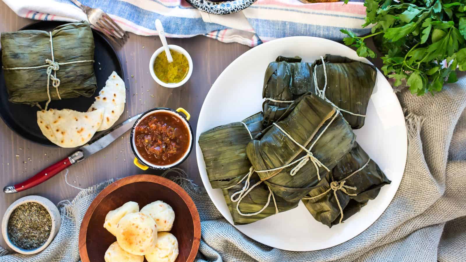 A plate of wrapped leaf parcels sits on a table with flatbreads, arepas, salsa, herbs, and condiments. Cloth napkins and utensils are also visible.
