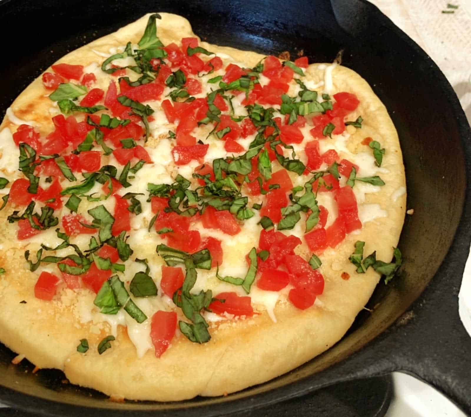 A cooked pizza in a cast iron skillet topped with chopped tomatoes, fresh basil, and melted cheese.