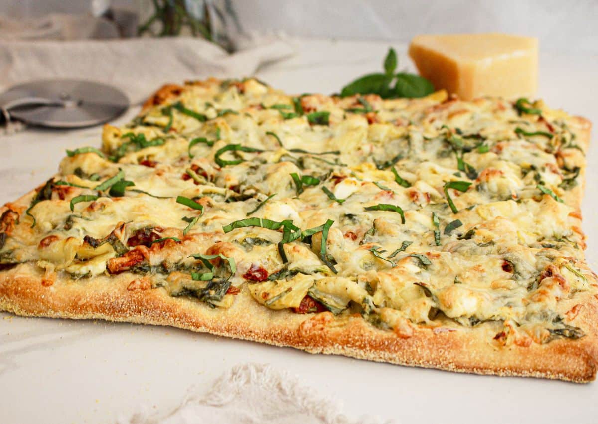 A rectangular flatbread pizza topped with cheese, spinach, artichokes, and basil sits on a white surface next to a pizza cutter and a block of cheese.
