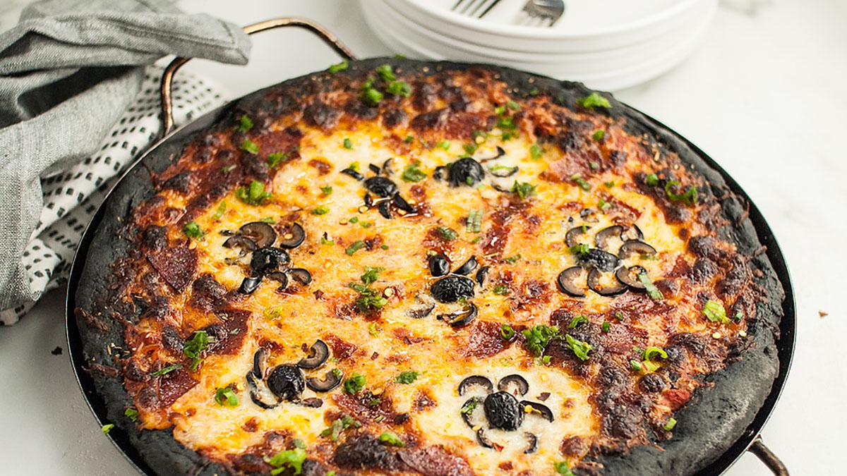 A round pizza with melted cheese, black olives, and herbs on a dark, crispy crust, served in a pan on a white surface with plates and forks in the background.