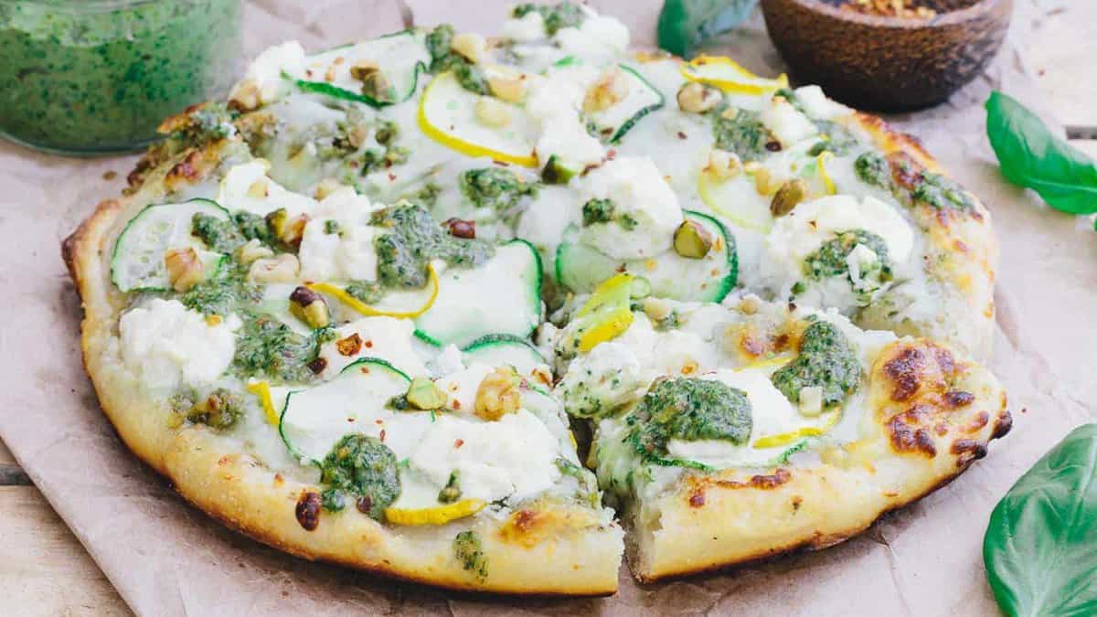 A vegetable pizza topped with zucchini slices, dollops of ricotta cheese, green pesto, and chopped nuts on parchment paper.