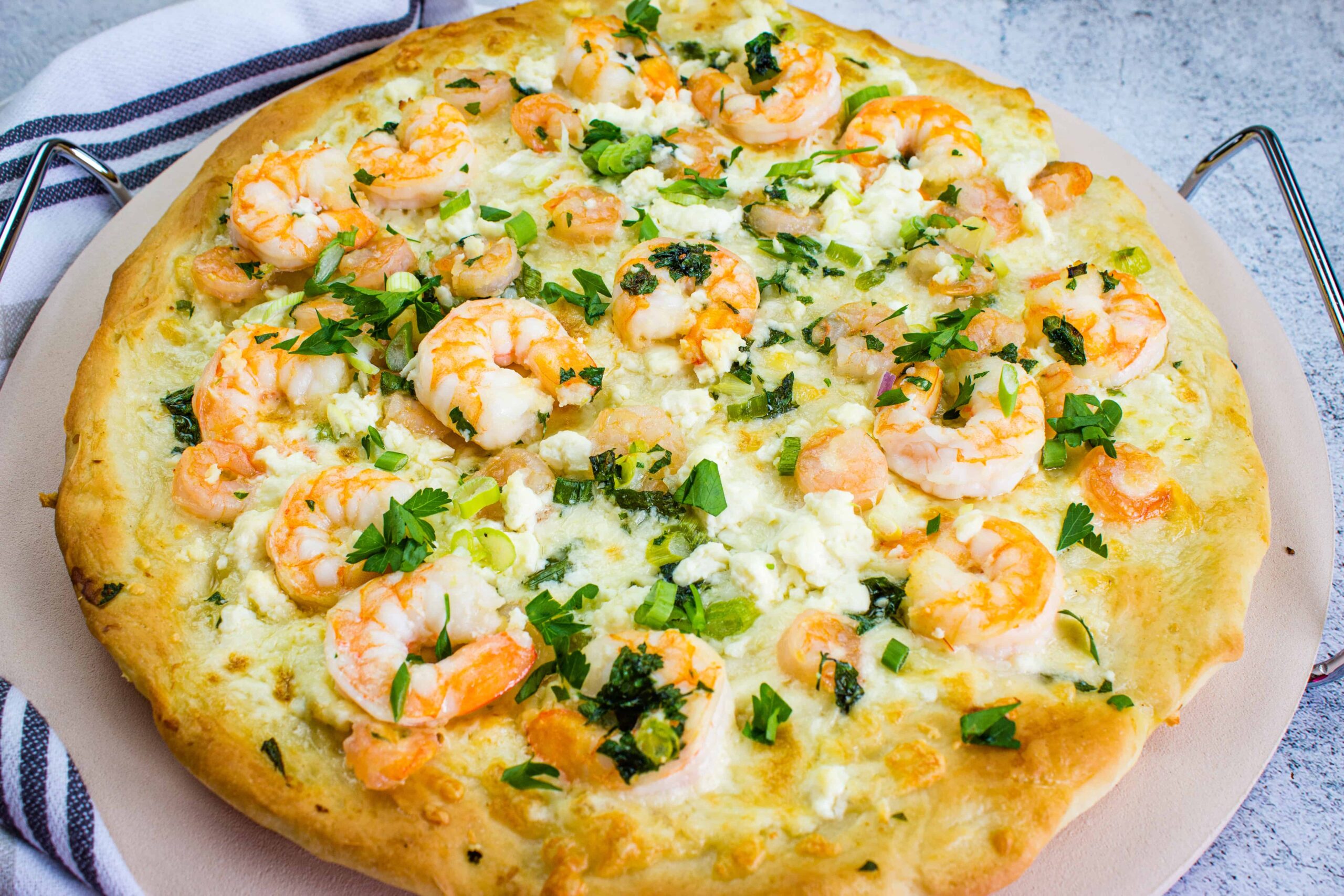 A baked pizza topped with shrimp, crumbled cheese, and chopped fresh herbs on a round pizza stone.