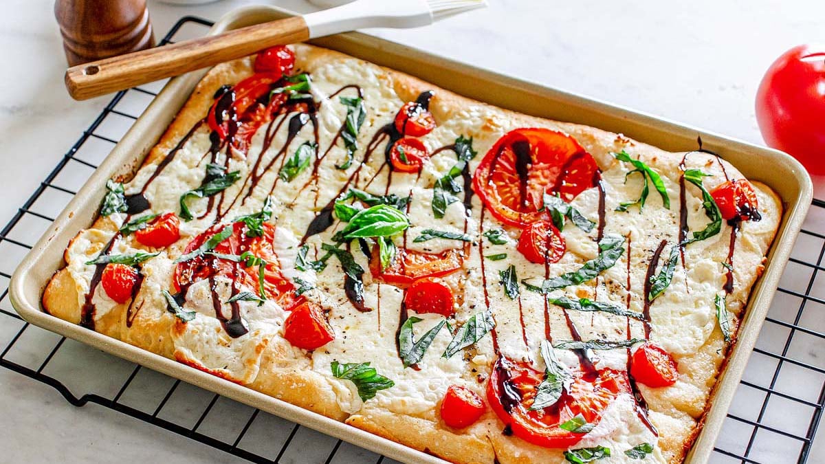 A baked rectangular focaccia topped with mozzarella, tomato slices, basil leaves, and a drizzle of balsamic glaze on a cooling rack.