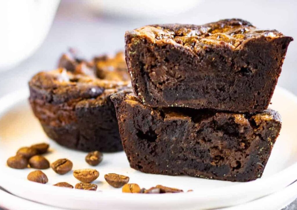 Two chocolate brownies stacked on a white plate, with whole coffee beans scattered beside them. Another brownie sits in the background.
