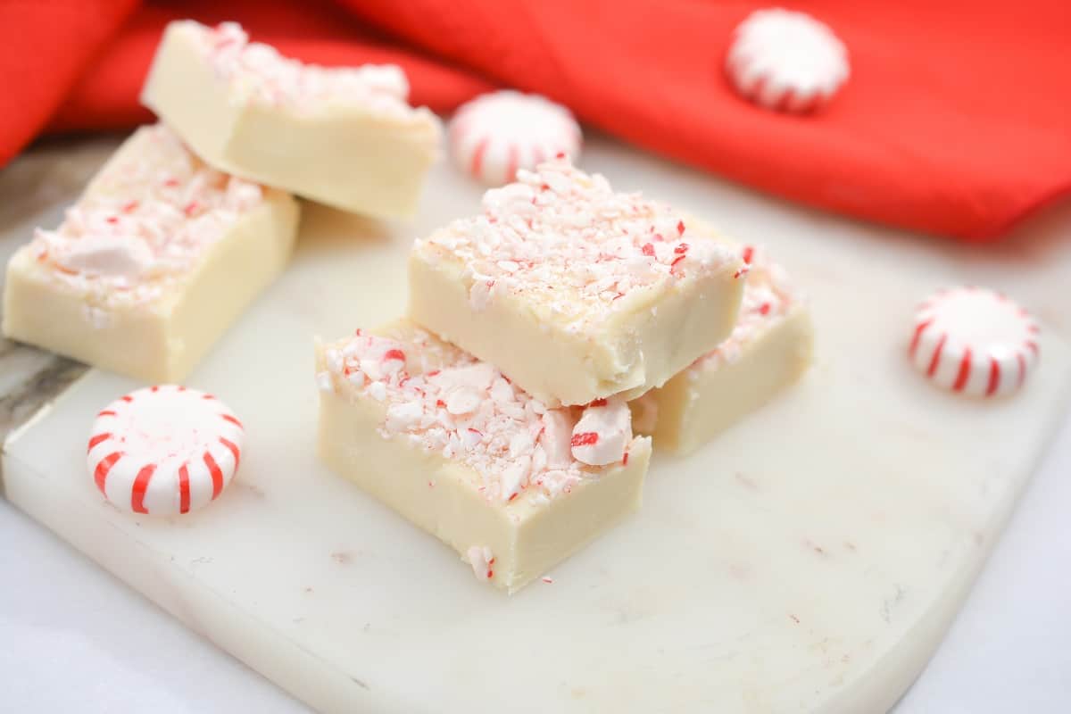 White chocolate peppermint fudge pieces topped with crushed peppermint, arranged on a marble surface with whole peppermint candies and a red cloth in the background.