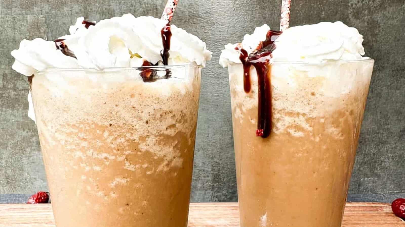 Two iced coffee drinks topped with whipped cream and chocolate syrup, each with a striped straw, sitting on a wooden surface against a gray background.