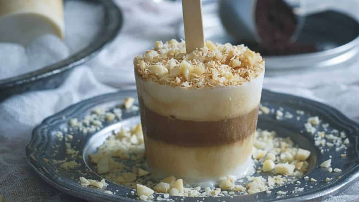 A layered ice cream popsicle with a wooden stick, topped with chopped nuts and coconut flakes, served on a metal plate with scattered nut pieces.