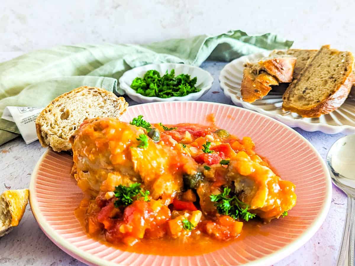 A plate of chicken cacciatore topped with chopped parsley, served with slices of bread and a small dish of extra parsley on the side.