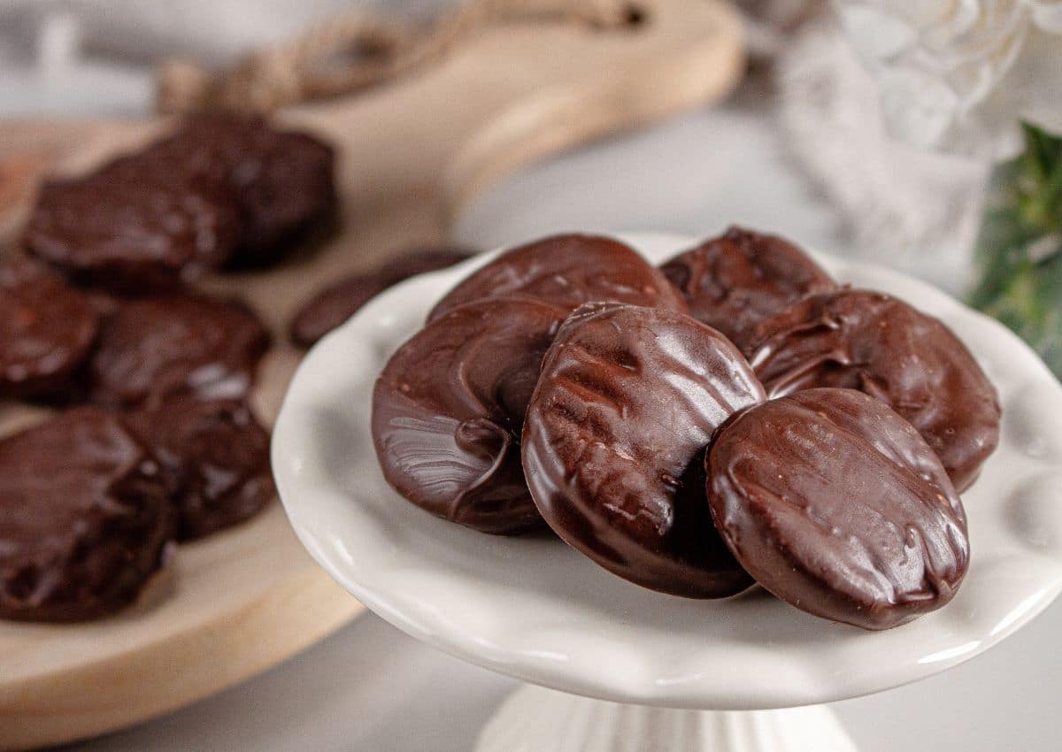 A white plate holds several round, chocolate-covered cookies, with more cookies visible on a wooden board in the background.