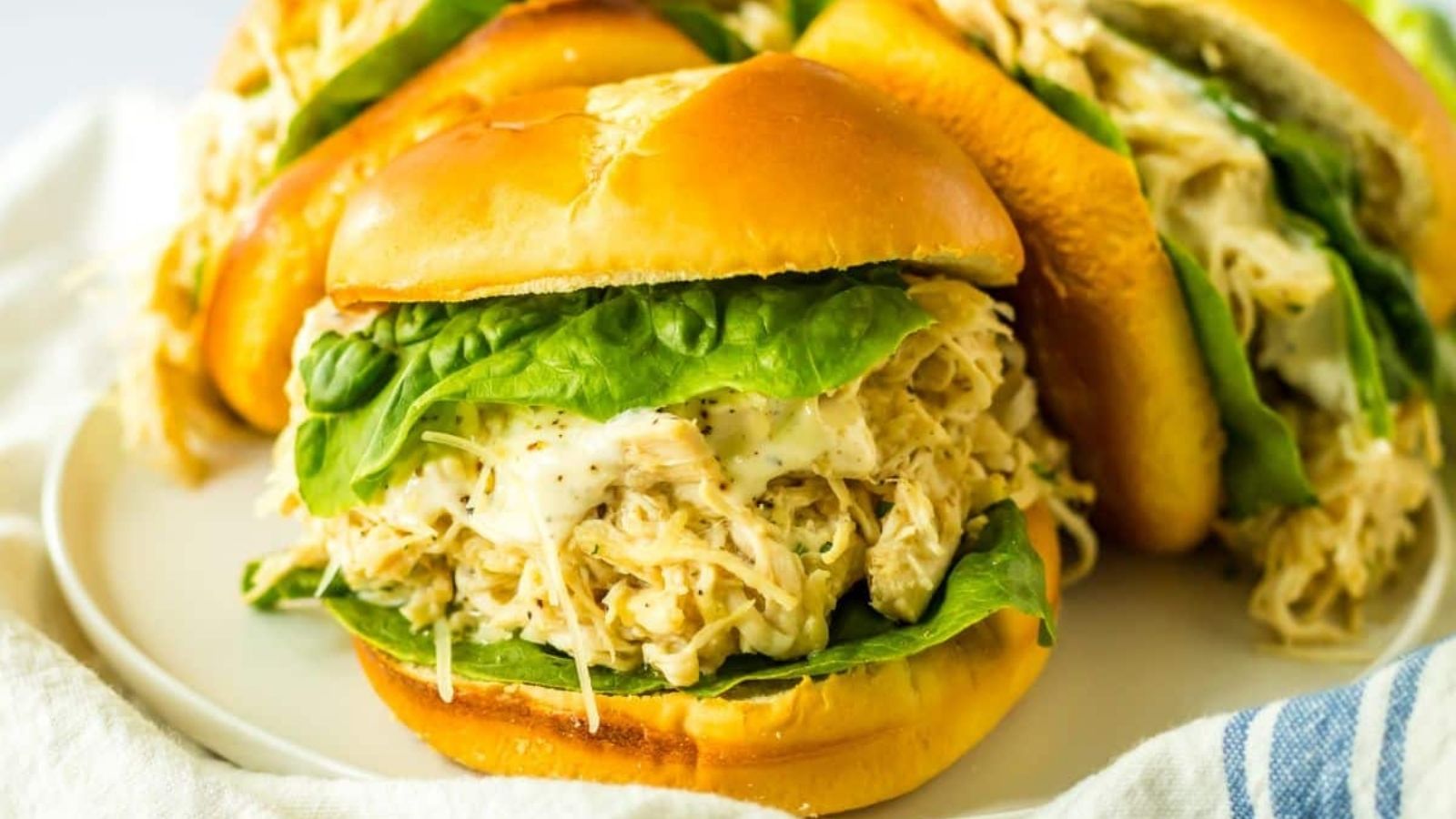 A shredded chicken sandwich with lettuce on a toasted bun, served on a white plate with similar sandwiches in the background.
