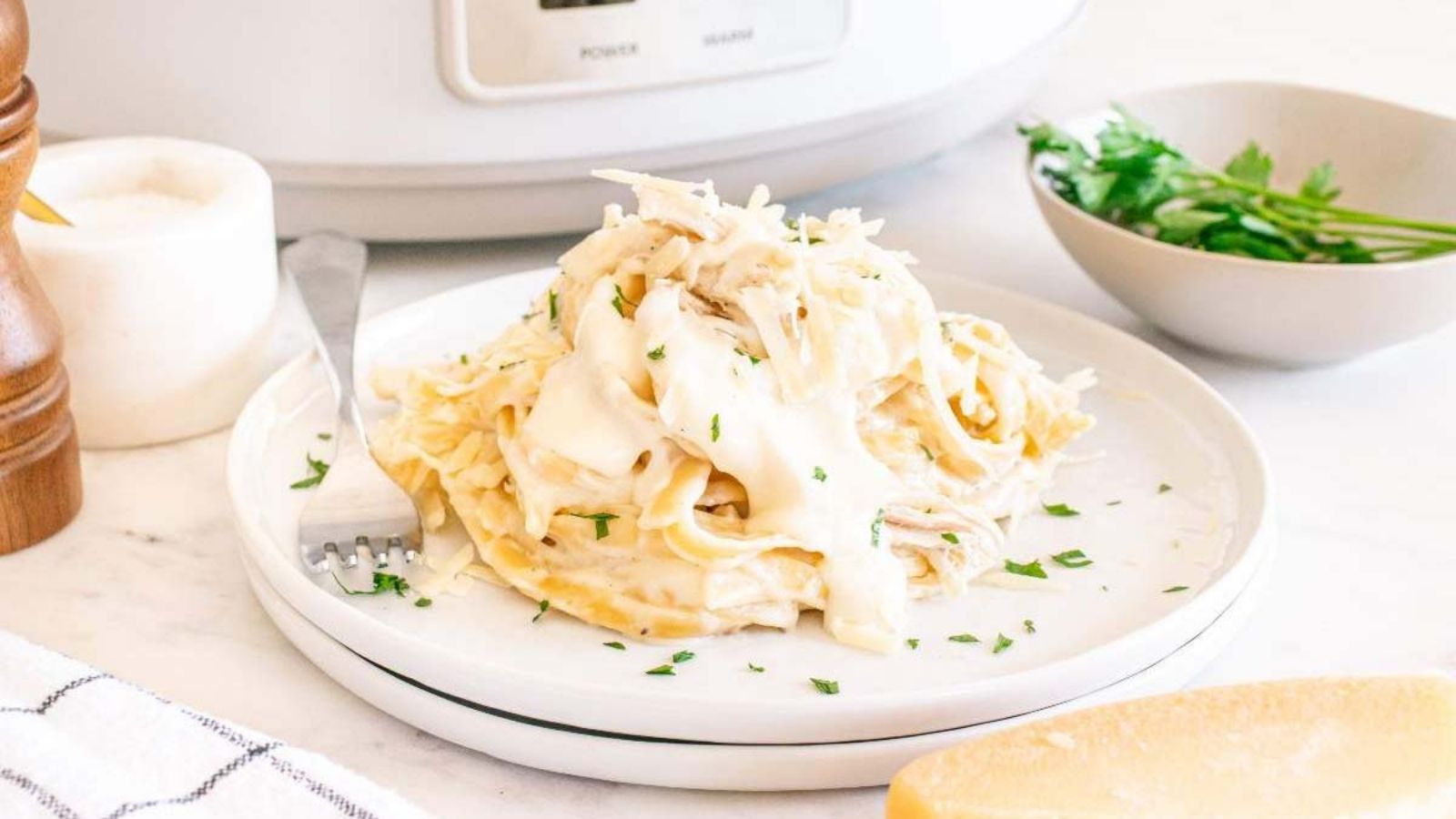 A plate of fettuccine Alfredo with shredded chicken, garnished with parsley, sits on a white plate beside a fork and a bowl of fresh herbs.