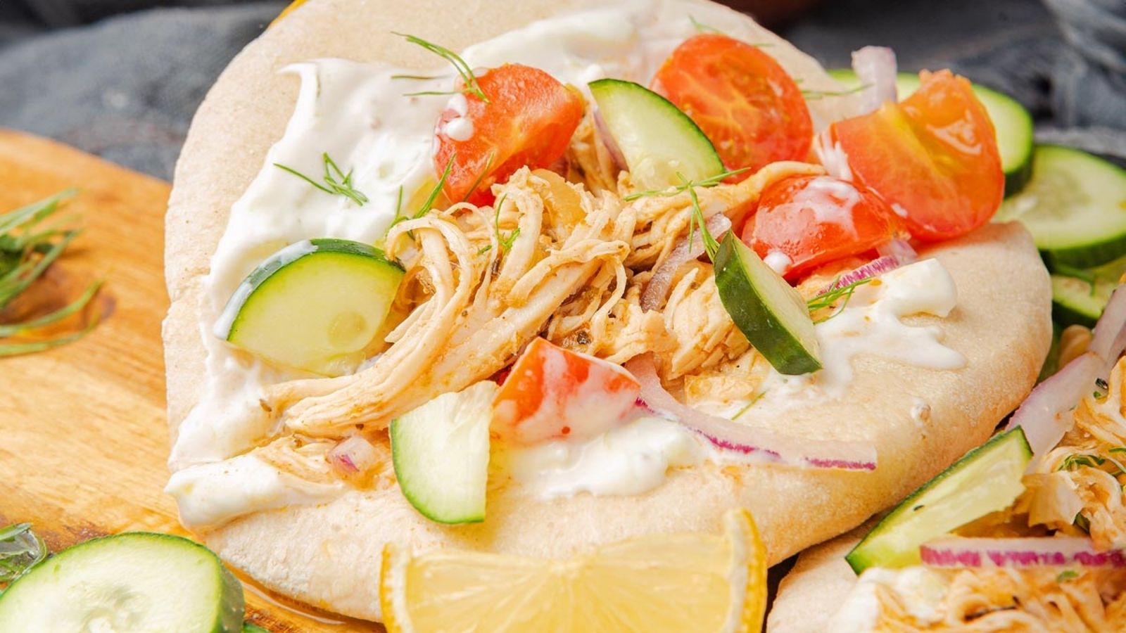 Flatbread topped with shredded chicken, sliced cucumbers, cherry tomatoes, red onions, and white sauce, with a lemon wedge on the side.