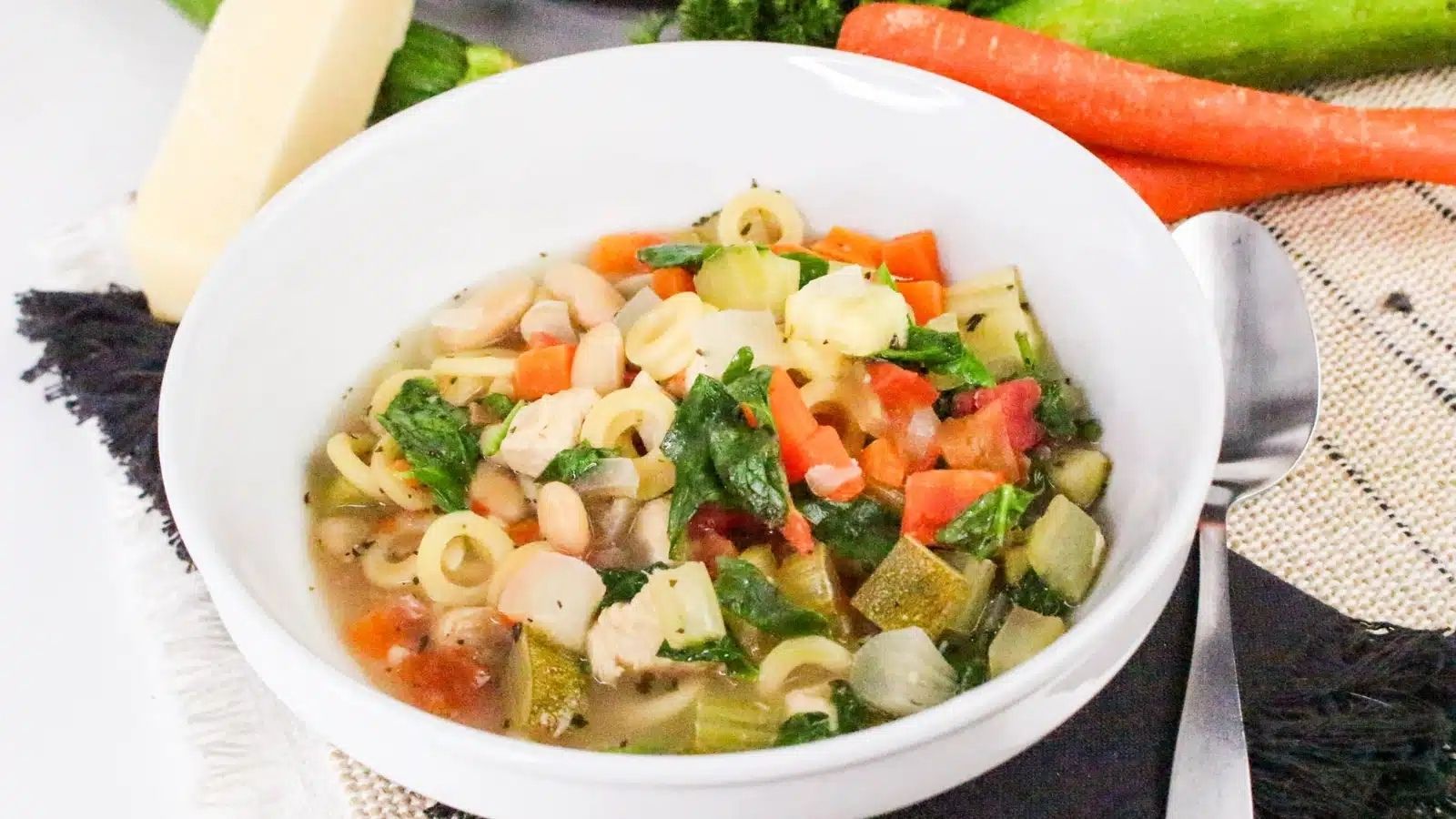 A white bowl filled with vegetable soup containing pasta, beans, spinach, carrots, zucchini, and herbs, placed on a placemat with a spoon beside it.