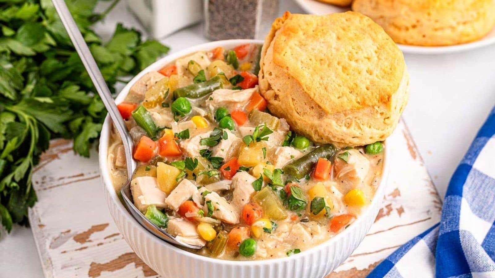 A bowl of chicken and vegetable stew topped with a biscuit, served on a rustic white surface with parsley and a blue checkered napkin nearby.