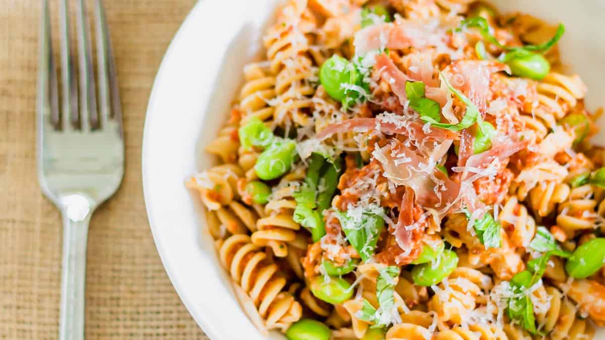 A white bowl of fusilli pasta topped with shredded cheese, green edamame, prosciutto, and fresh herbs sits next to a metal fork on a textured surface.
