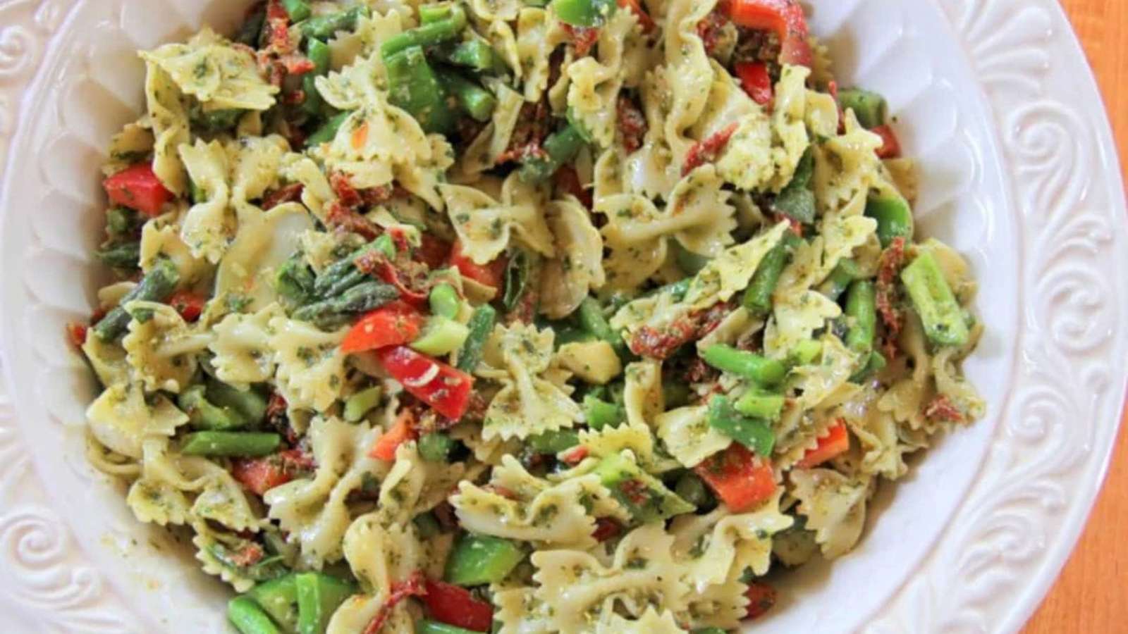 A bowl of bowtie pasta mixed with green beans, red bell peppers, sun-dried tomatoes, and a green pesto sauce.