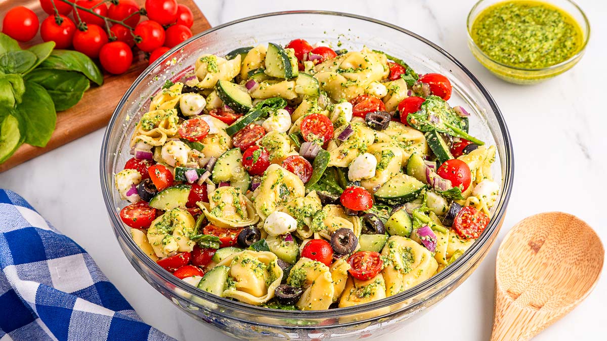 A glass bowl filled with tortellini pasta salad, mixed with cherry tomatoes, cucumbers, mozzarella balls, olives, red onion, and pesto. A small bowl of pesto and fresh vegetables are nearby.