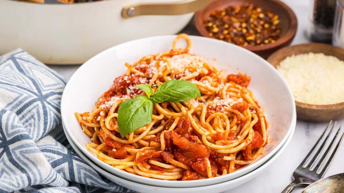 A bowl of spaghetti with tomato sauce, grated cheese, and basil leaves, placed on a table with a napkin, fork, and bowls of grated cheese and red pepper flakes.