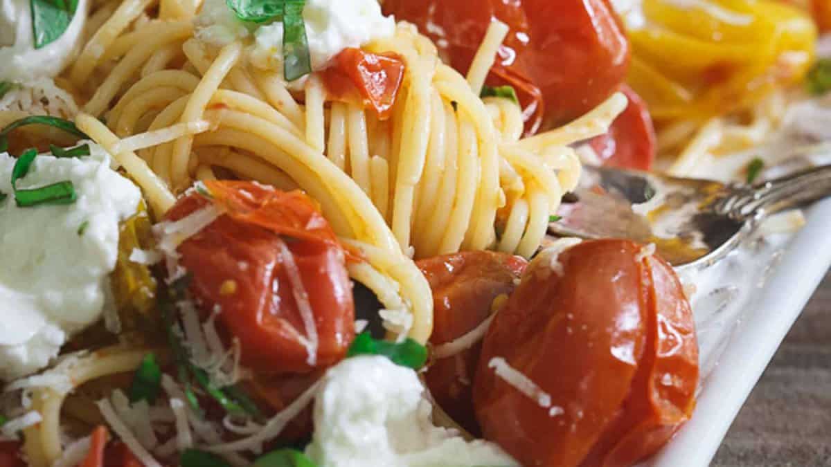 Close-up of spaghetti with roasted cherry tomatoes, fresh basil, dollops of ricotta cheese, and a fork on a white plate.