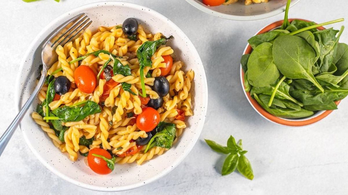 A bowl of fusilli pasta with cherry tomatoes, black olives, spinach, and basil. A small bowl of fresh spinach is next to it. A fork rests in the pasta bowl.