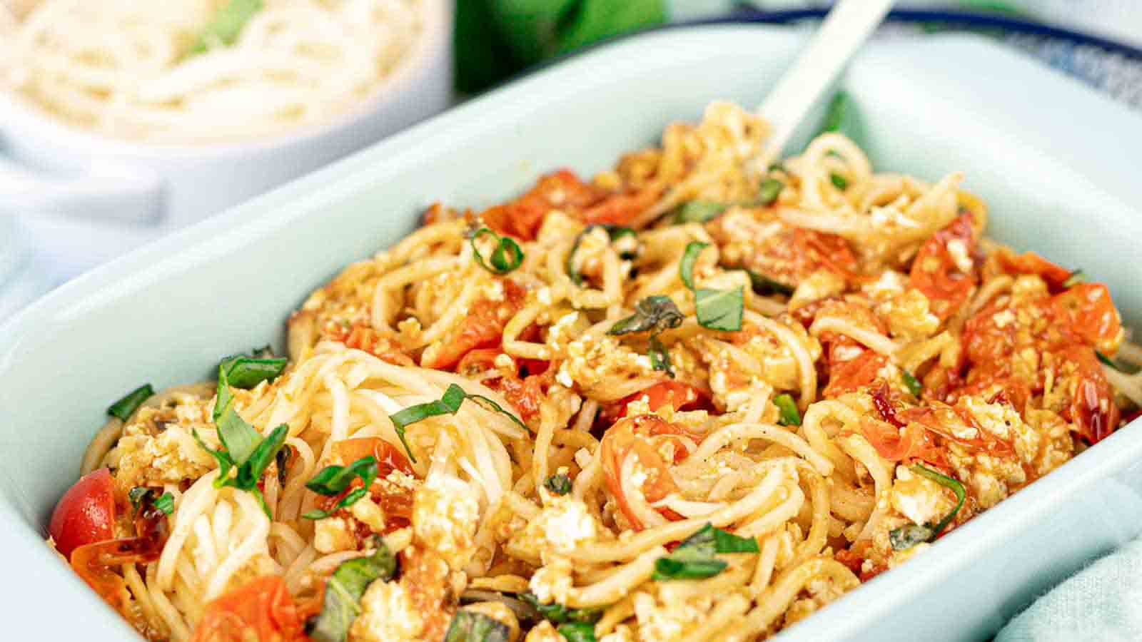 A casserole dish filled with baked spaghetti, cherry tomatoes, crumbled cheese, and fresh basil, with a white serving utensil.