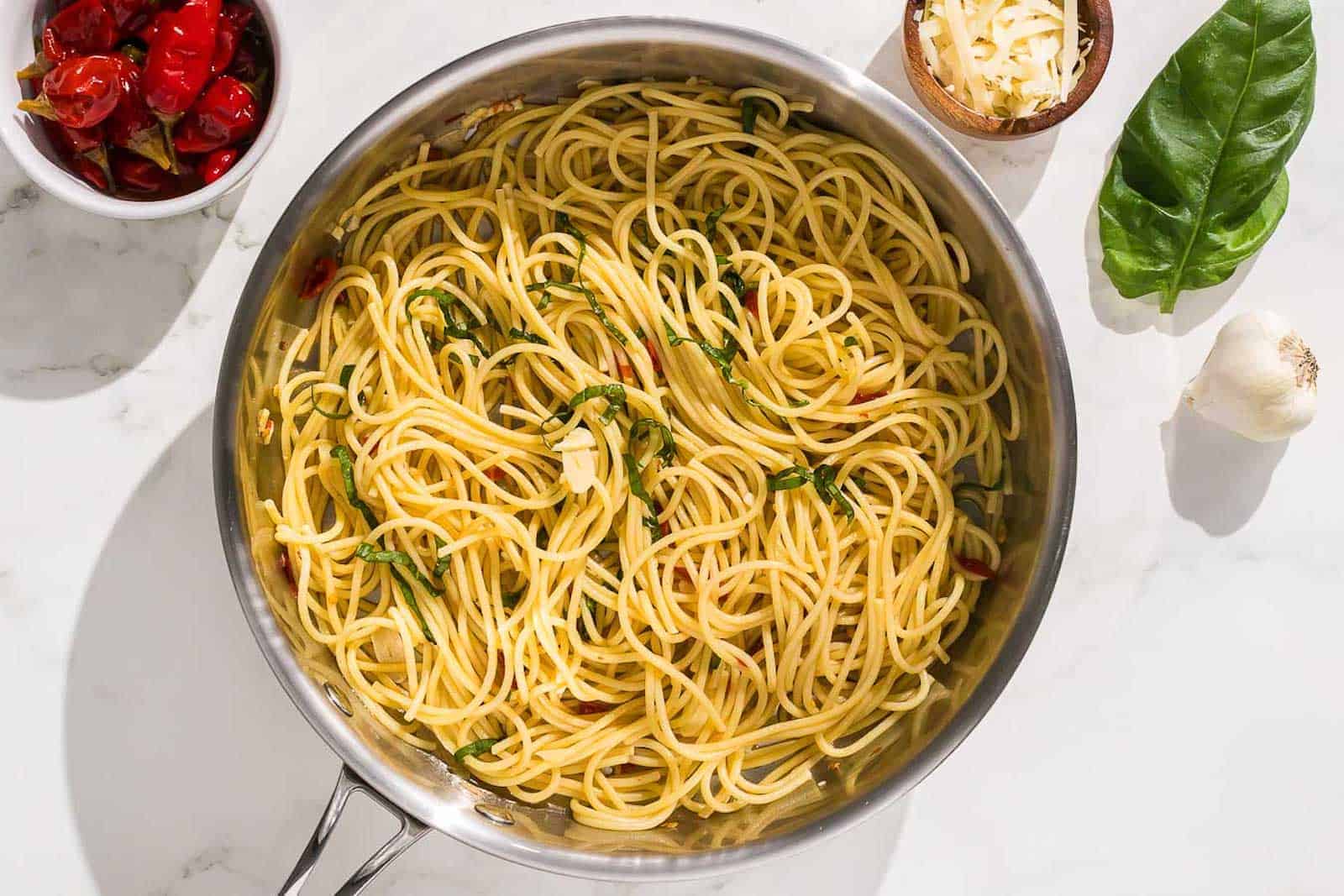 A pan of cooked spaghetti with garlic and herbs, surrounded by a bowl of roasted tomatoes, grated cheese, basil leaves, and a garlic bulb on a white surface.