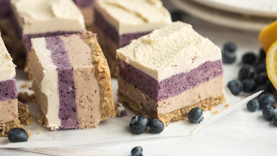 Squares of layered blueberry cheesecake with a crumbly crust, some blueberries scattered around, and lemon slices partially visible on the side.