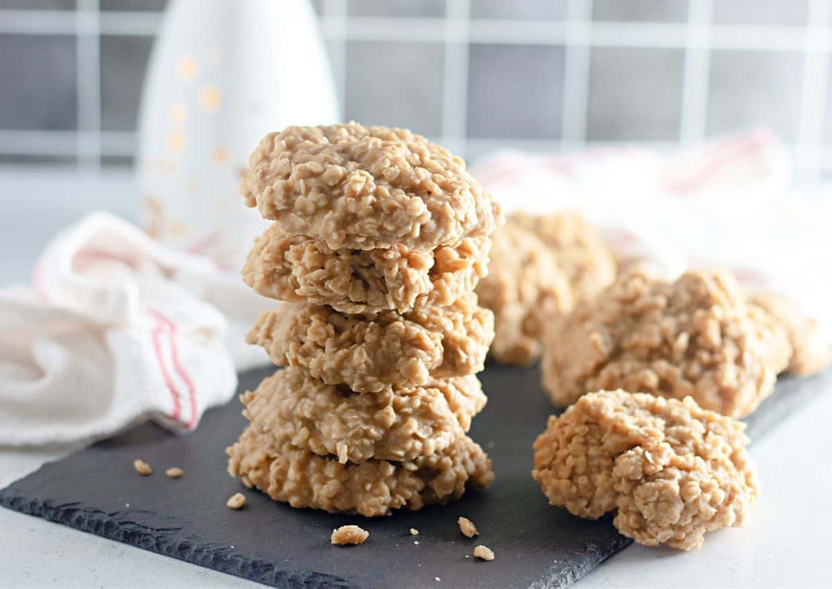 A stack of five no-bake oatmeal cookies on a dark slate surface, with more cookies and a white towel in the background.