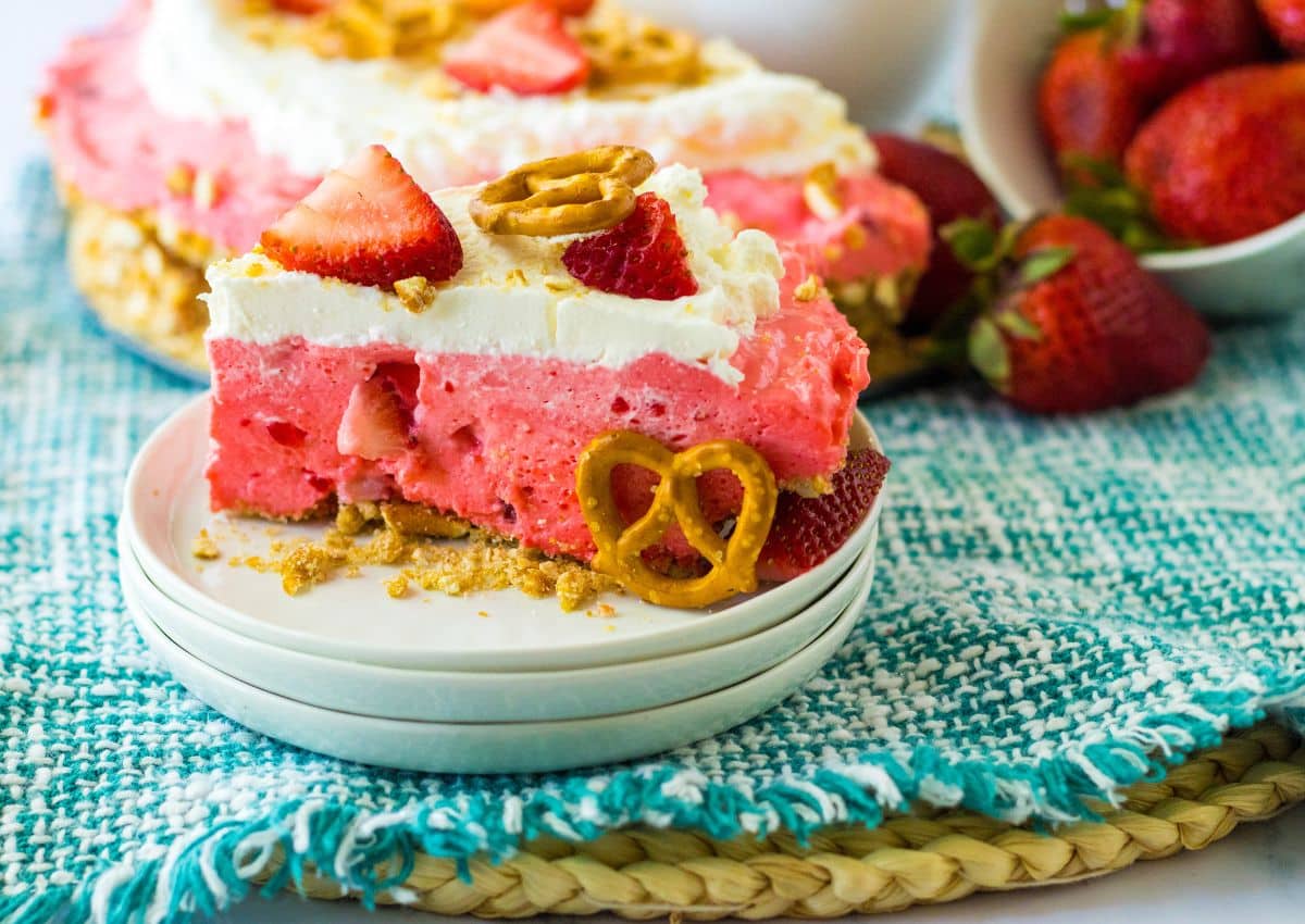 A slice of strawberry dessert with whipped topping, fresh strawberries, and pretzels on a white plate, set on a blue woven mat with whole strawberries in the background.