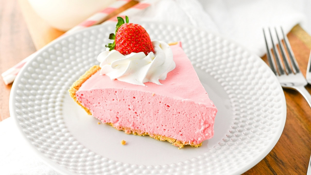 A slice of pink strawberry mousse pie in a graham cracker crust, topped with whipped cream and a whole strawberry, on a white plate.