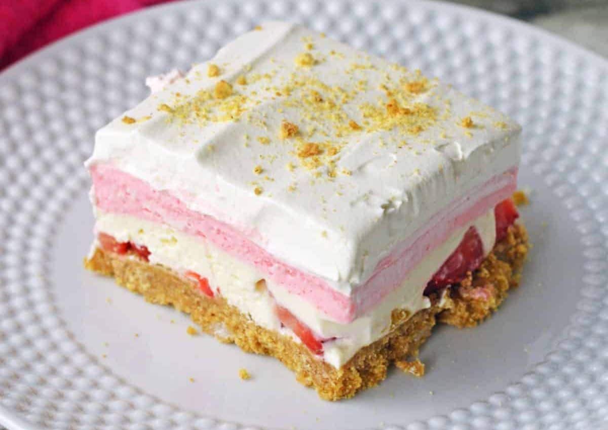A dessert slice with a crumbly base, creamy white and pink layers, a layer of sliced strawberries, and whipped topping, served on a white plate.