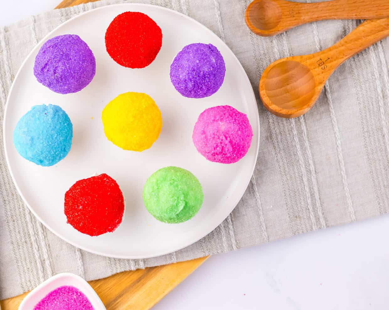 A white plate with nine colorful sugar-coated balls arranged in a circle, next to three wooden measuring spoons on a striped cloth.
