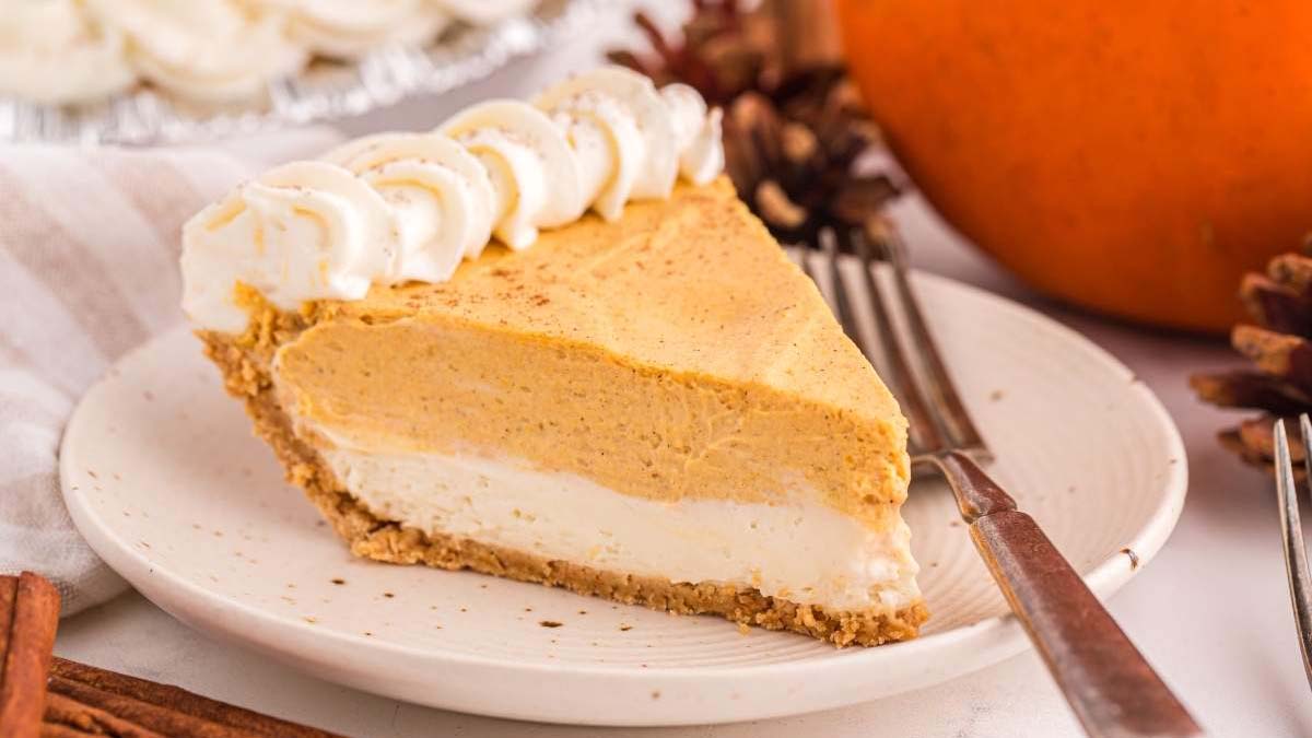A slice of layered pumpkin cream pie with whipped topping sits on a white plate, next to a fork and autumn-themed decorations.