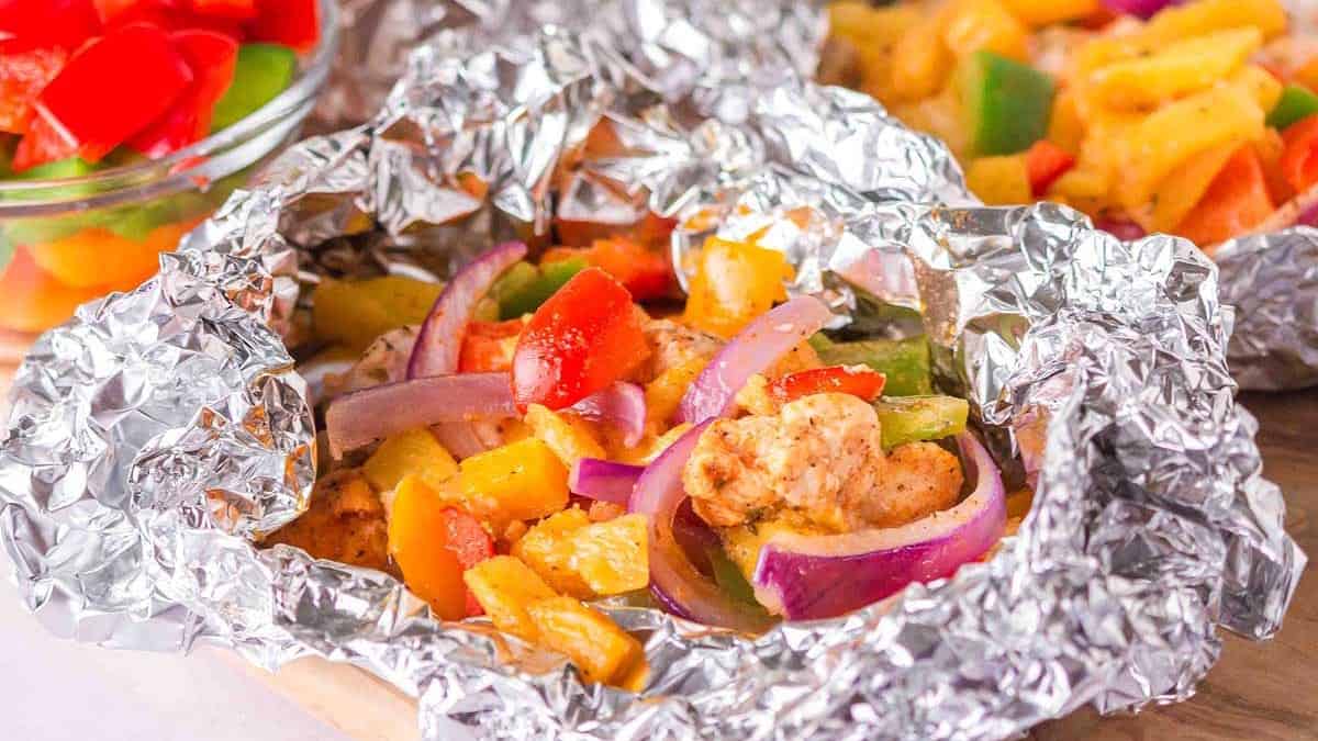 Chopped chicken, bell peppers, and red onions cooked in an open foil packet on a wooden surface.
