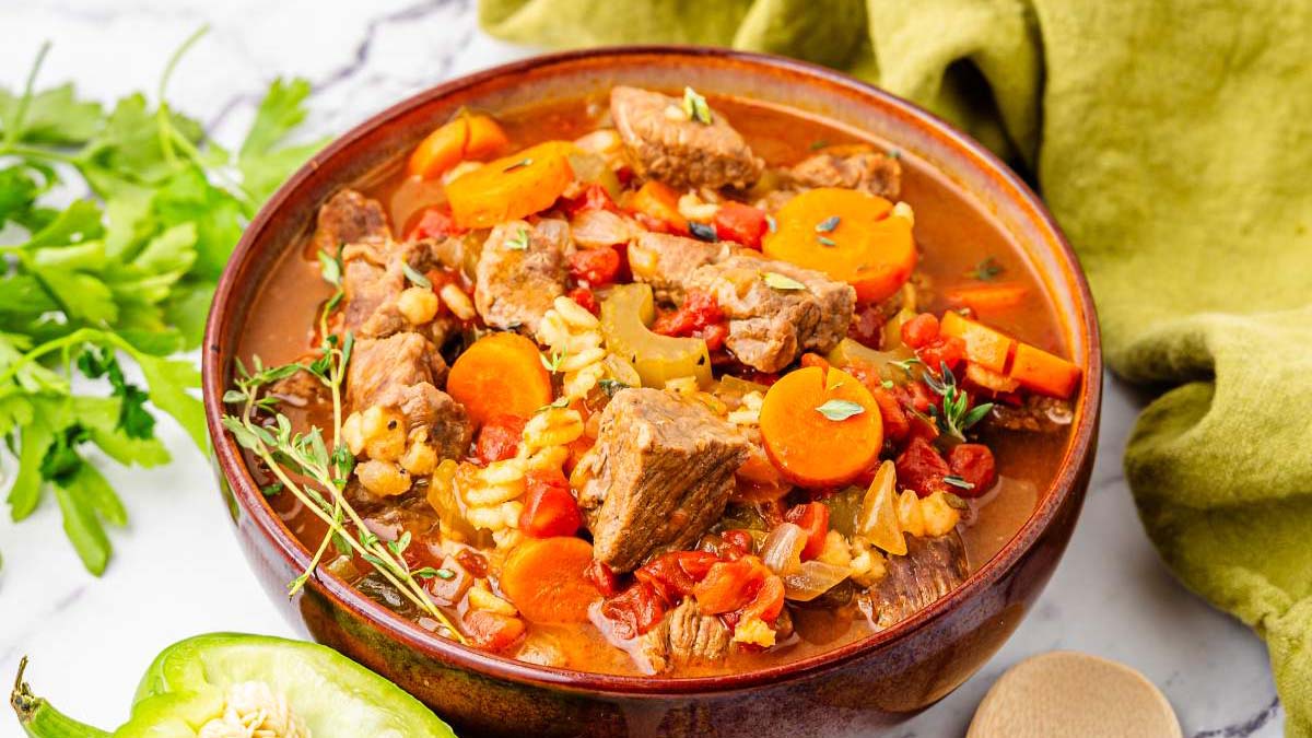 A bowl of beef and vegetable stew with carrots, tomatoes, and herbs, placed on a white surface next to parsley and a green bell pepper.